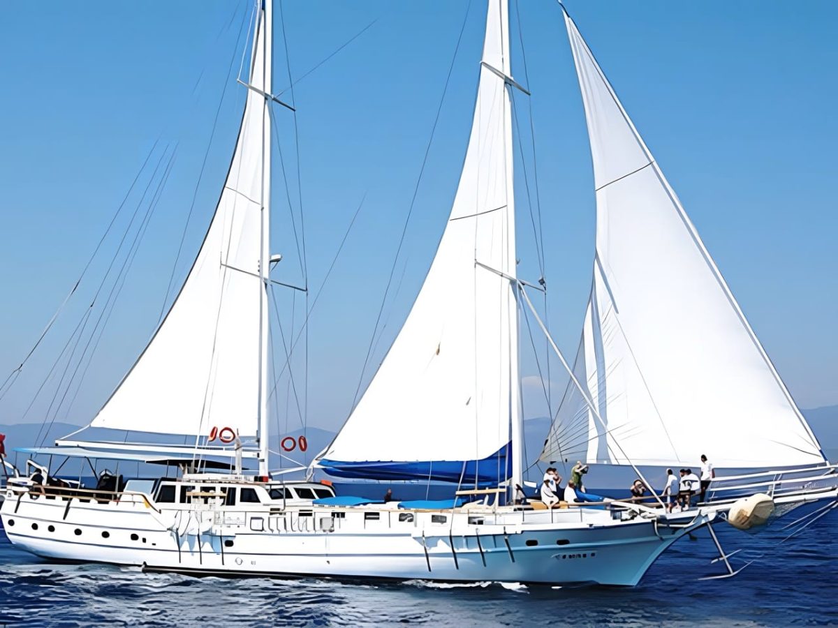 Cemre IV under sail with guests on the foredeck and the sea dark blue beneath a clear sky