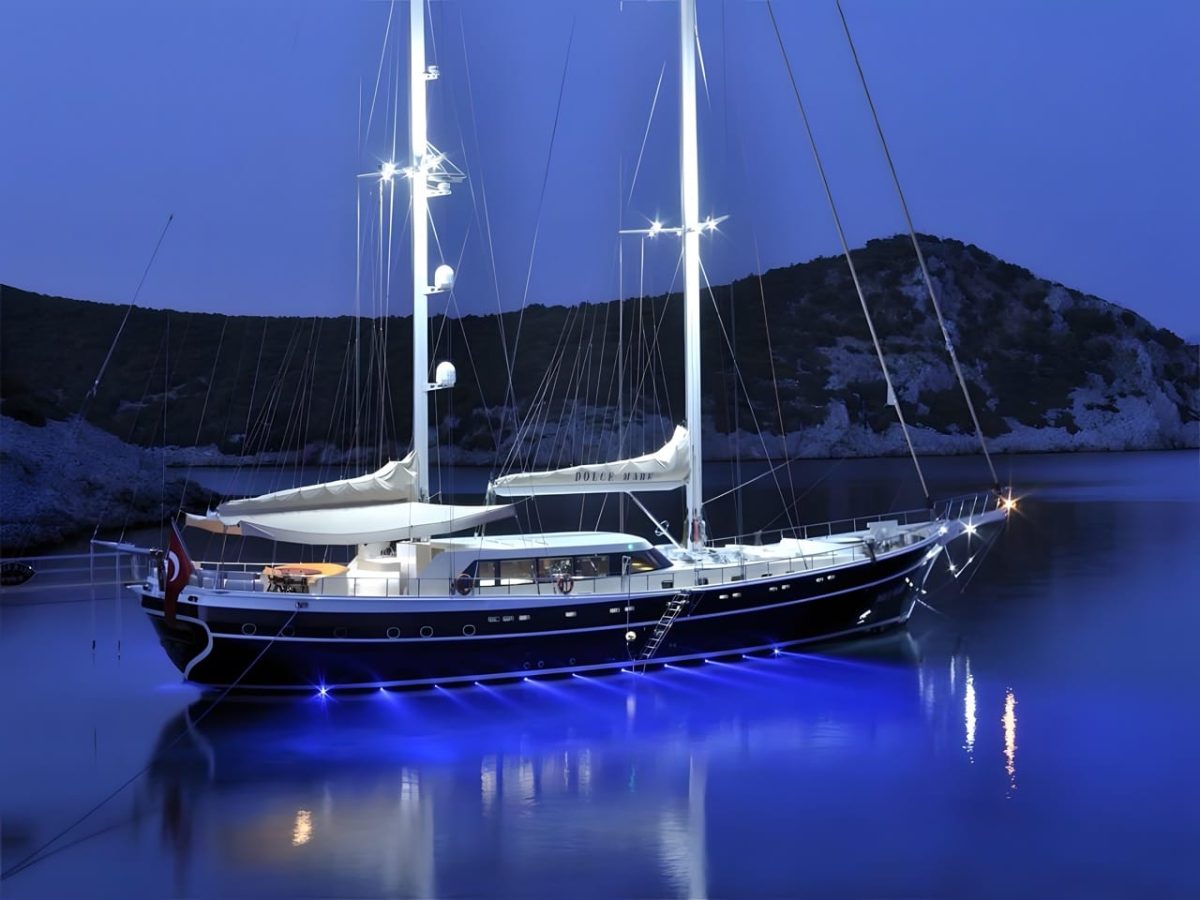 Dolce Mare moored at dusk, her dark hull lit from below and her masts picked out in white against a rocky headland