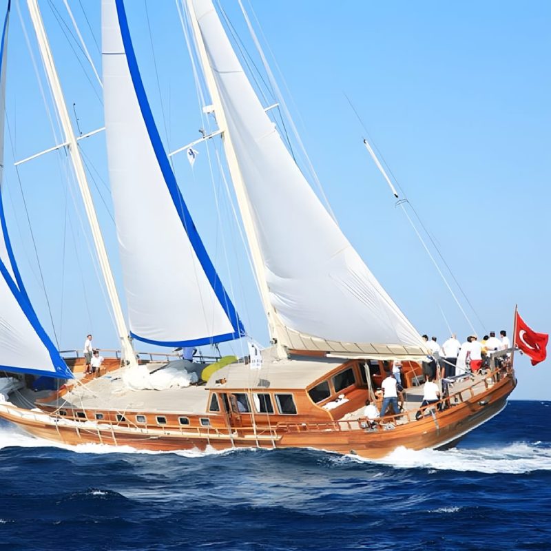 Kaya Guneri III heeling under blue and white sails, crew working along her varnished deck and the Turkish flag streaming aft
