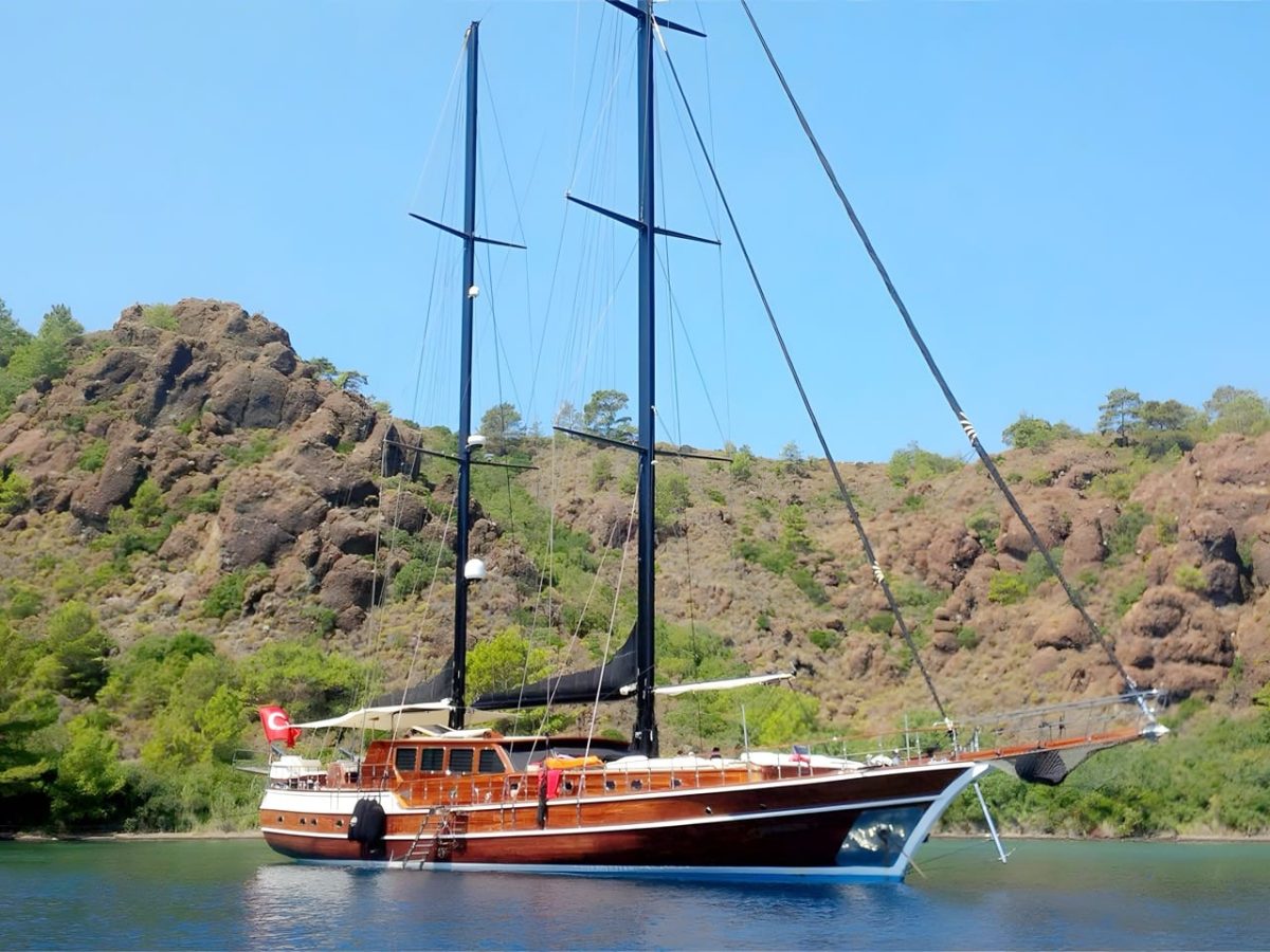 Kaya Guneri Plus at anchor with her varnished mahogany hull and black masts against a red rock hillside