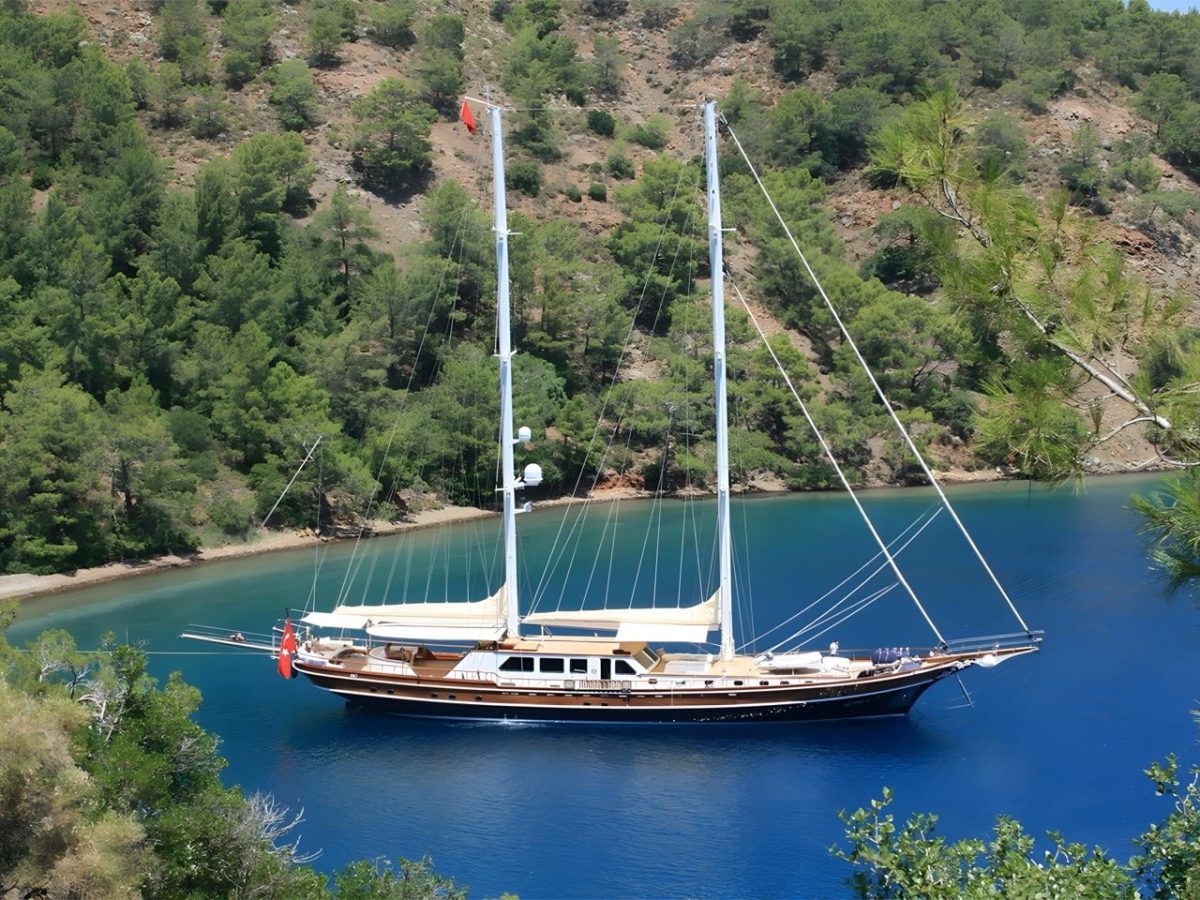 The gulet Kaya Guneri V at anchor in a narrow pine-walled inlet, her two masts bare and the water deep blue around her