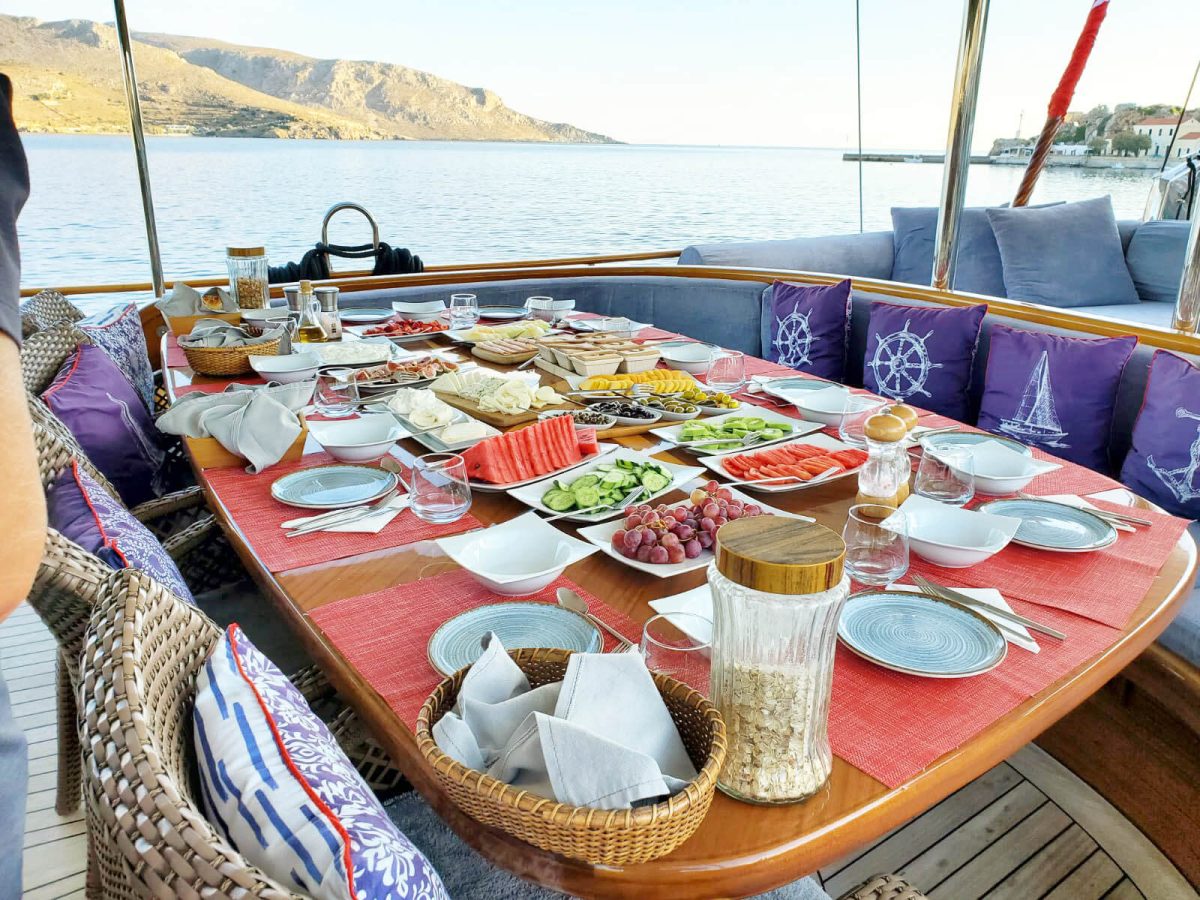 A generous breakfast spread of cheese, fruit and olives laid on the aft deck table, with a harbour town in the distance.