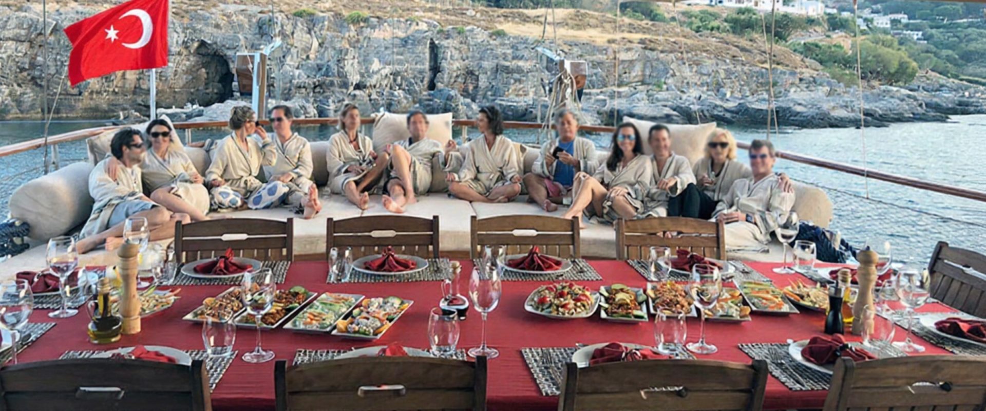 Guests in robes at a long table set for lunch on the aft deck of a chartered gulet, anchored in a cove on the Turkish coast.