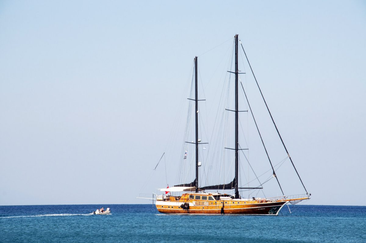 A traditional two-masted gulet at anchor in clear blue water, sails furled, with the tender approaching.