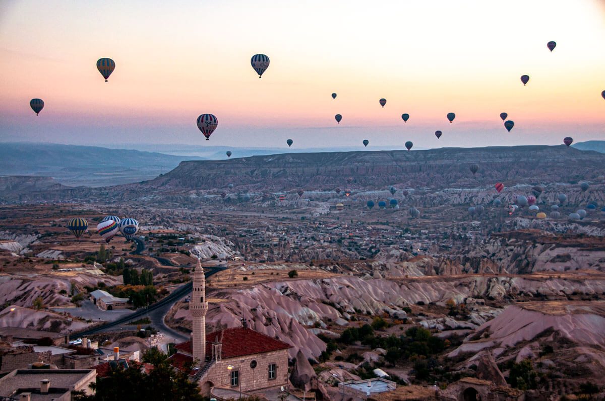 Dozens of hot air balloons over the valleys of Cappadocia at sunrise, a village minaret in the foreground