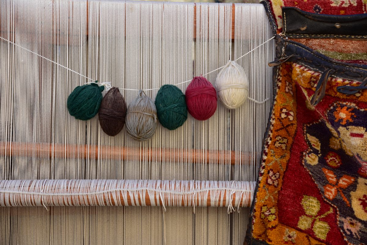 Balls of dyed wool strung across the warp of a carpet loom, a finished rug beside it